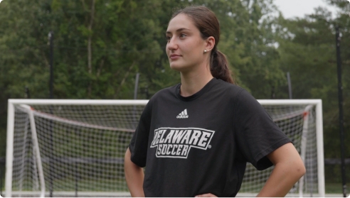 Video thumbnail of a young woman in a Delaware Soccer shirt standing on a field in front of a goal.