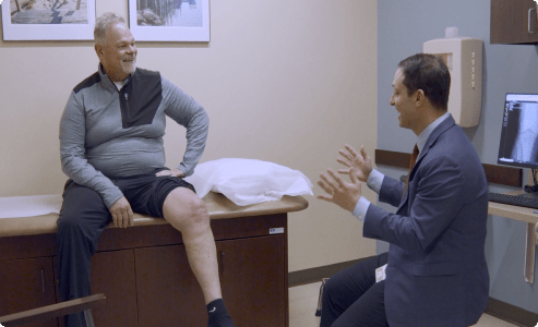 A patient with short gray hair sitting on an exam table, talking with a physician.