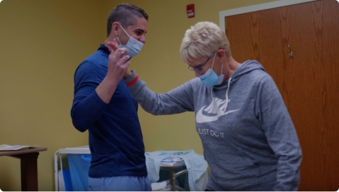 Video thumbnail showing a masked healthcare provider assisting a patient in a rehabilitation setting.